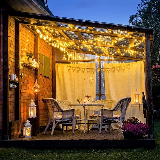 View over cozy outdoor terrace with table and chairs, very romantic lighting, white lanterns, candles burning, led string party lights and bulbs with star effect, in the evening.